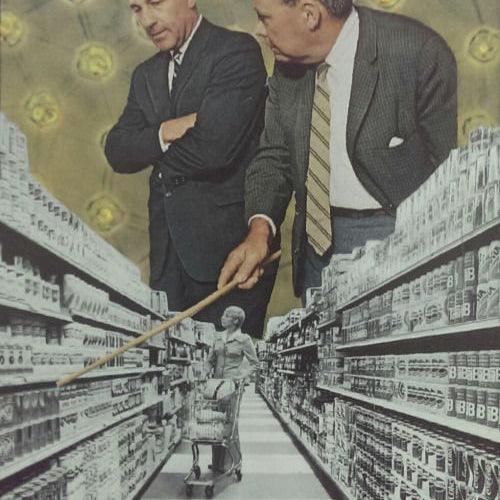 Two large men in suits stand above a supermarket aisle, with one pointing at shelves using a stick, while a woman shops below.