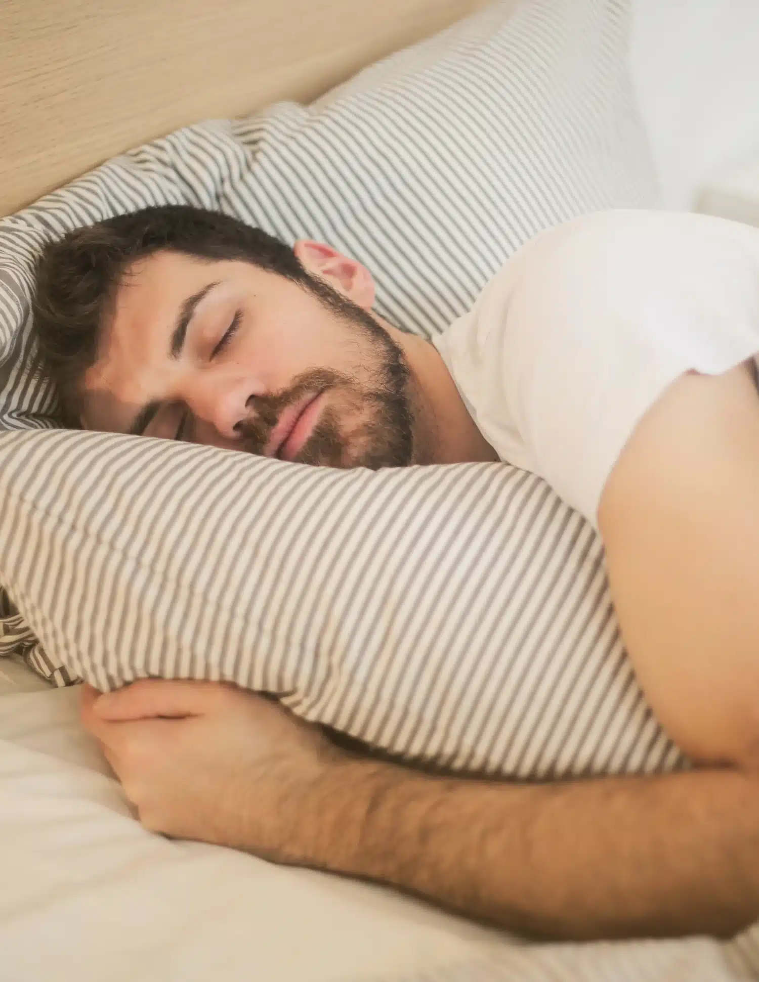 A man with dark hair and a beard sleeps soundly on a striped pillow, possibly enjoying the calming effects of CBN, wearing a white t-shirt and resting on light-colored bedding.