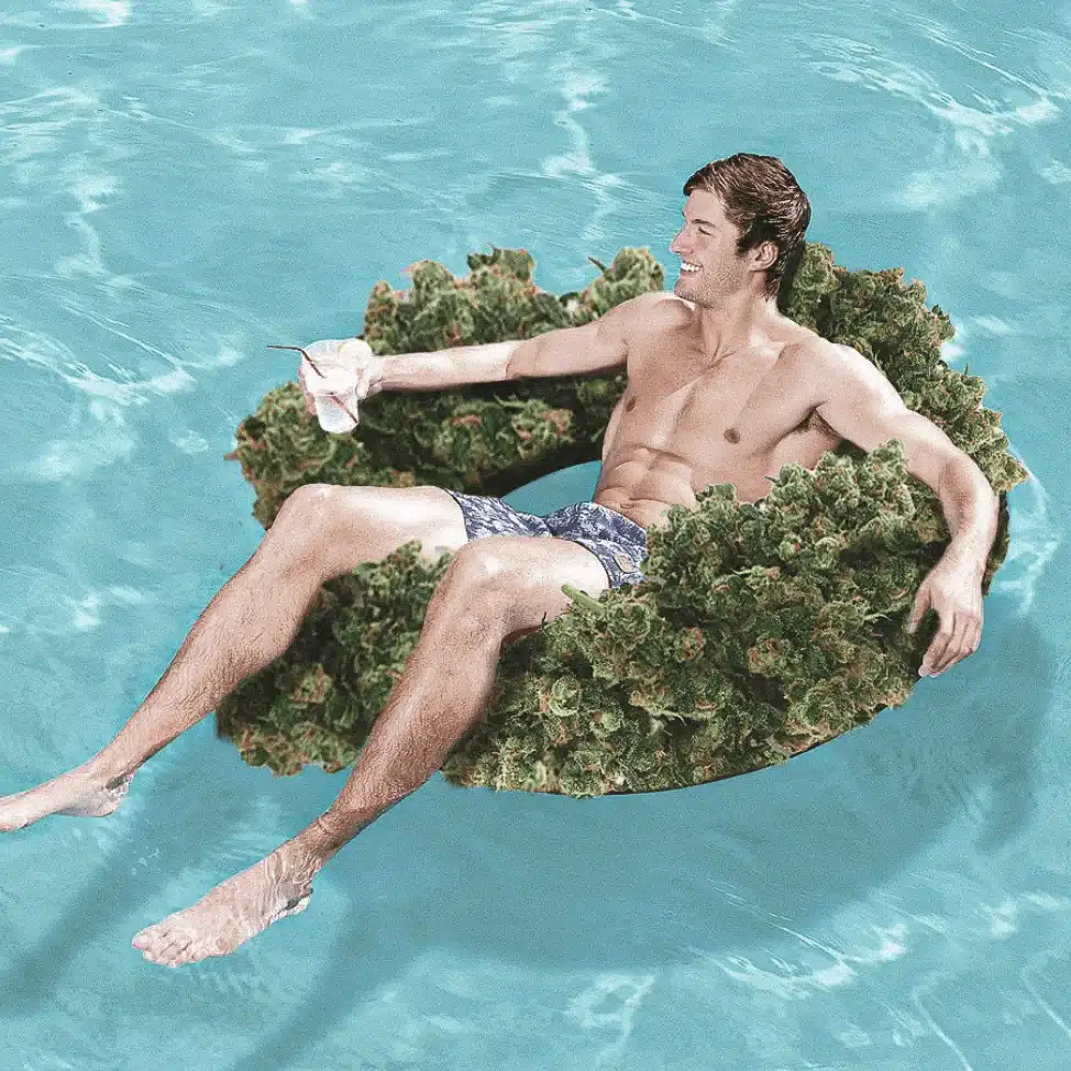 A man in swim trunks relaxes in a swimming pool on an inner tube made of green plant material, holding a drink and smiling.