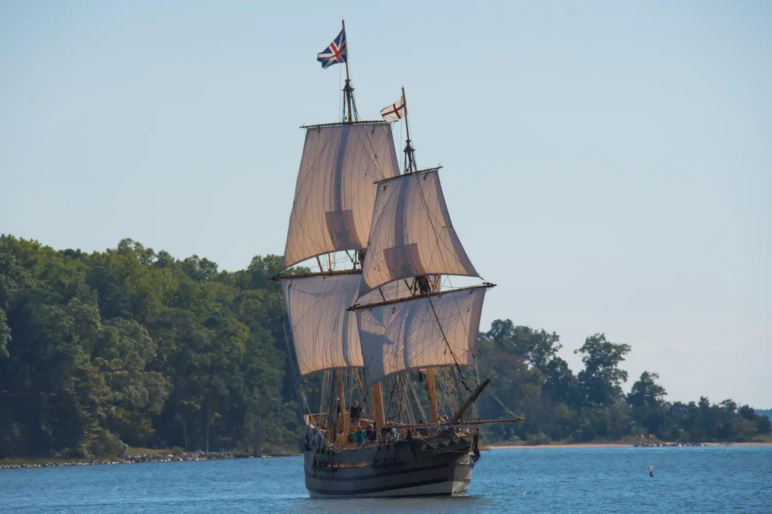 A large wooden sailing ship with three masts and British flags sails on a calm river near a forested shoreline.