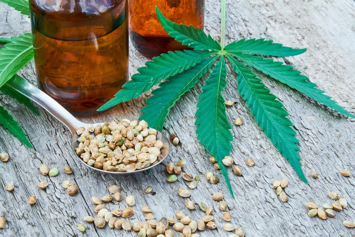 A spoonful of hemp seeds, two amber glass bottles, and a cannabis leaf on a wooden surface.