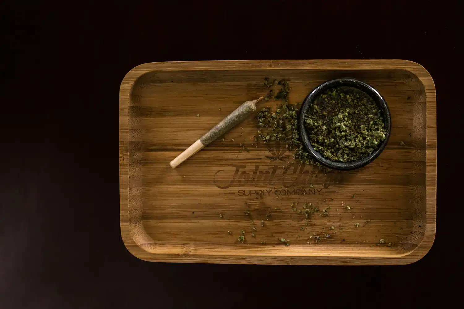 A rolled joint and a small bowl of loose cannabis sit on a wooden tray with scattered cannabis pieces.