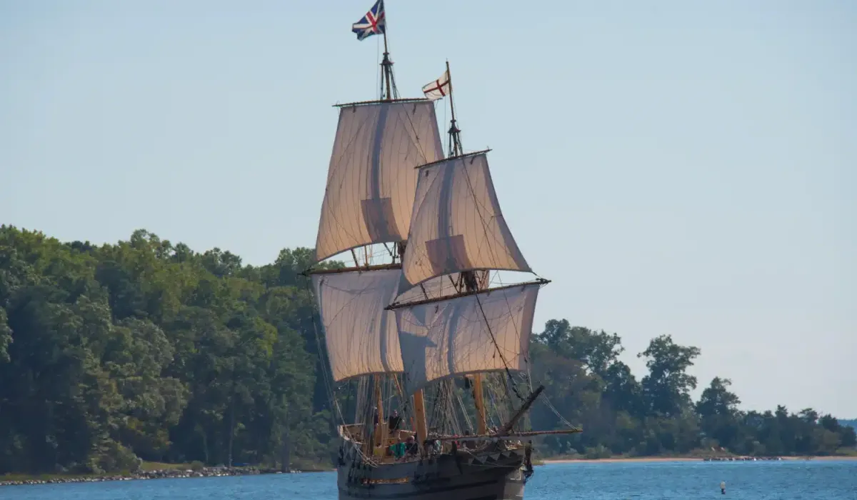 A large wooden sailing ship with three masts and British flags sails on a calm river near a forested shoreline.