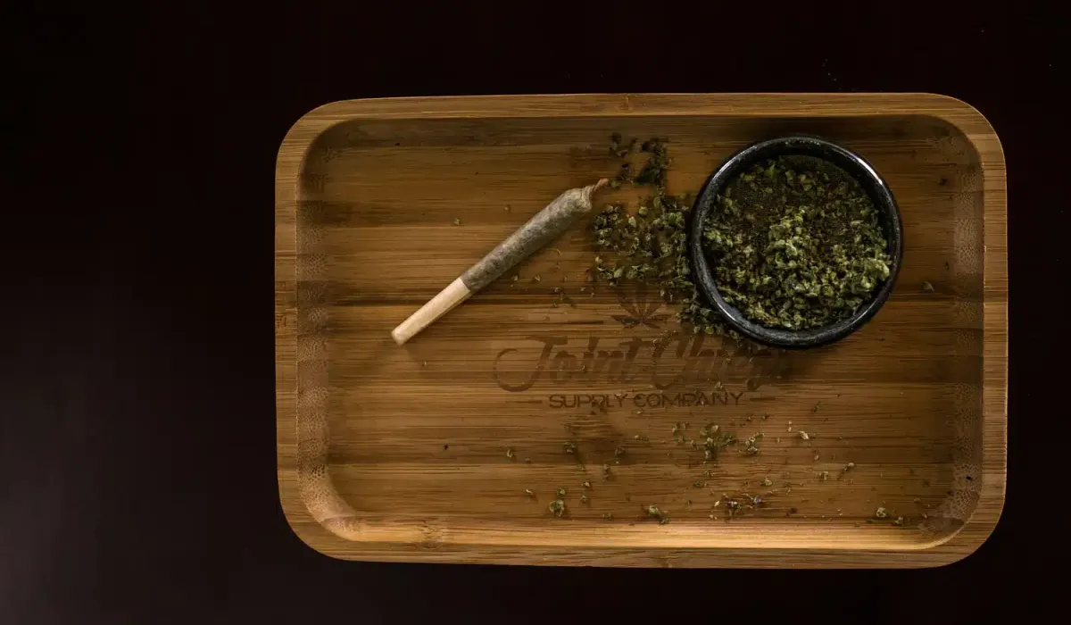 A rolled joint and a small bowl of loose cannabis sit on a wooden tray with scattered cannabis pieces.
