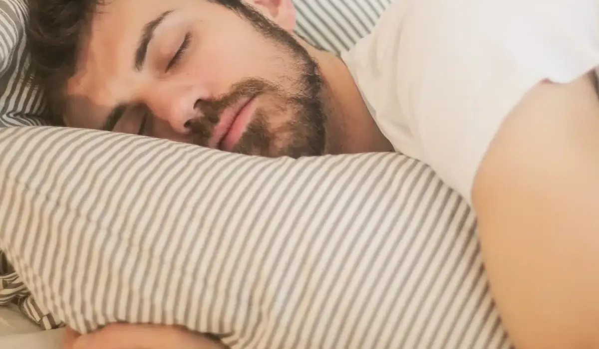 A man with dark hair and a beard sleeps soundly on a striped pillow, possibly enjoying the calming effects of CBN, wearing a white t-shirt and resting on light-colored bedding.