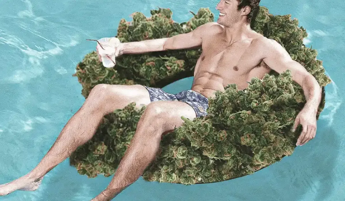 A man in swim trunks relaxes in a swimming pool on an inner tube made of green plant material, holding a drink and smiling.
