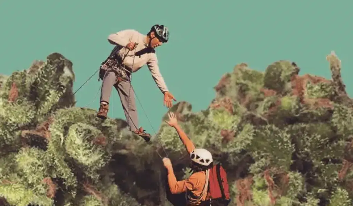 Two climbers in helmets and harnesses are on a rocky, moss-covered surface; one climber extends a hand to help the other up. The background is a plain teal color.