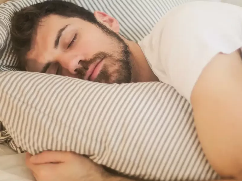 A man with dark hair and a beard sleeps soundly on a striped pillow, possibly enjoying the calming effects of CBN, wearing a white t-shirt and resting on light-colored bedding.