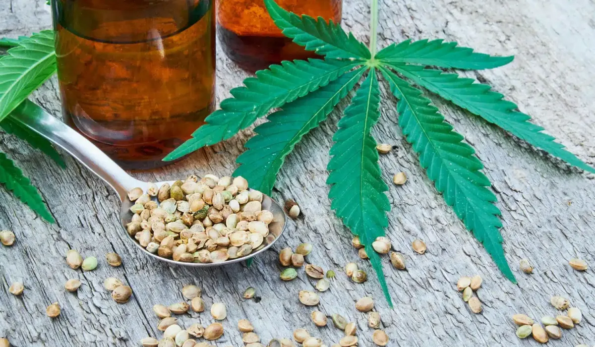 A spoonful of hemp seeds, two amber glass bottles, and a cannabis leaf on a wooden surface.