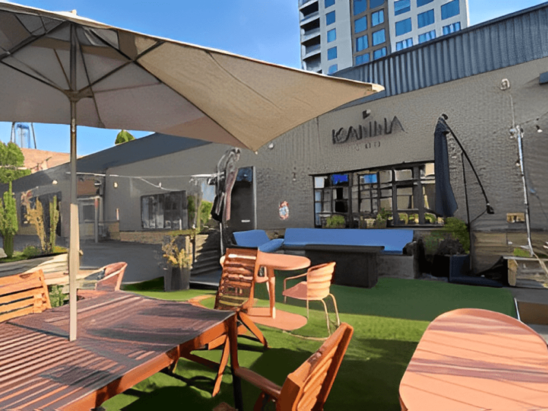 Outdoor seating area with wooden tables and chairs under umbrellas, artificial grass flooring, and a modern building labeled “KONA” in the background—contact Kanna CBD Store for more details.